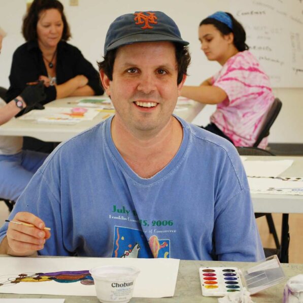 Scott wears a baseball hat and smiles while holding a paint brush in a CATA workshop