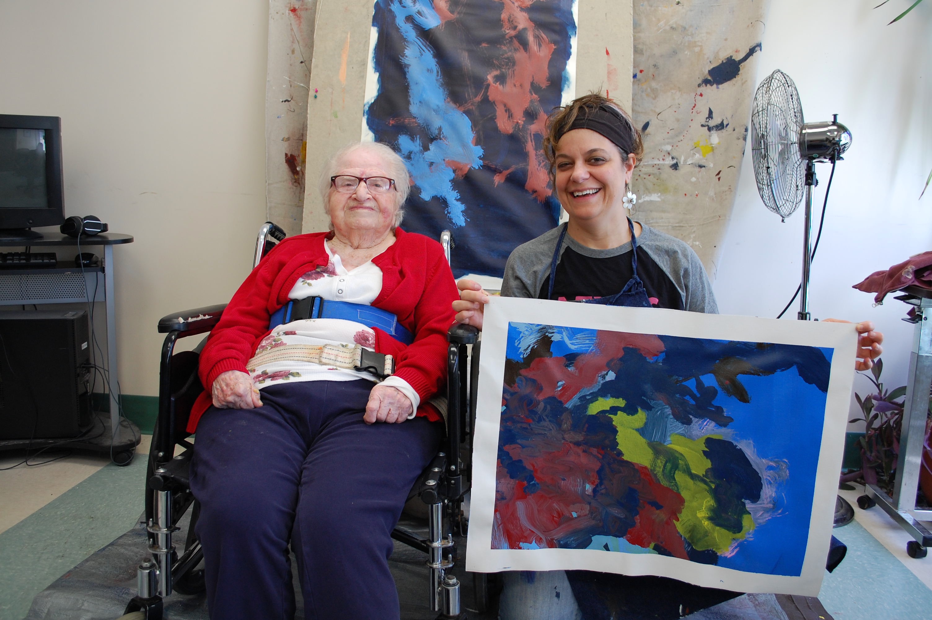 CATA artist Ruthie sits in a wheelchair next to Faculty Artist Stefanie, who holds Ruthie's new painting and smiles. The painting is abstract with broad strokes of red, dark blue, and yellow paint on top of a light blue background.