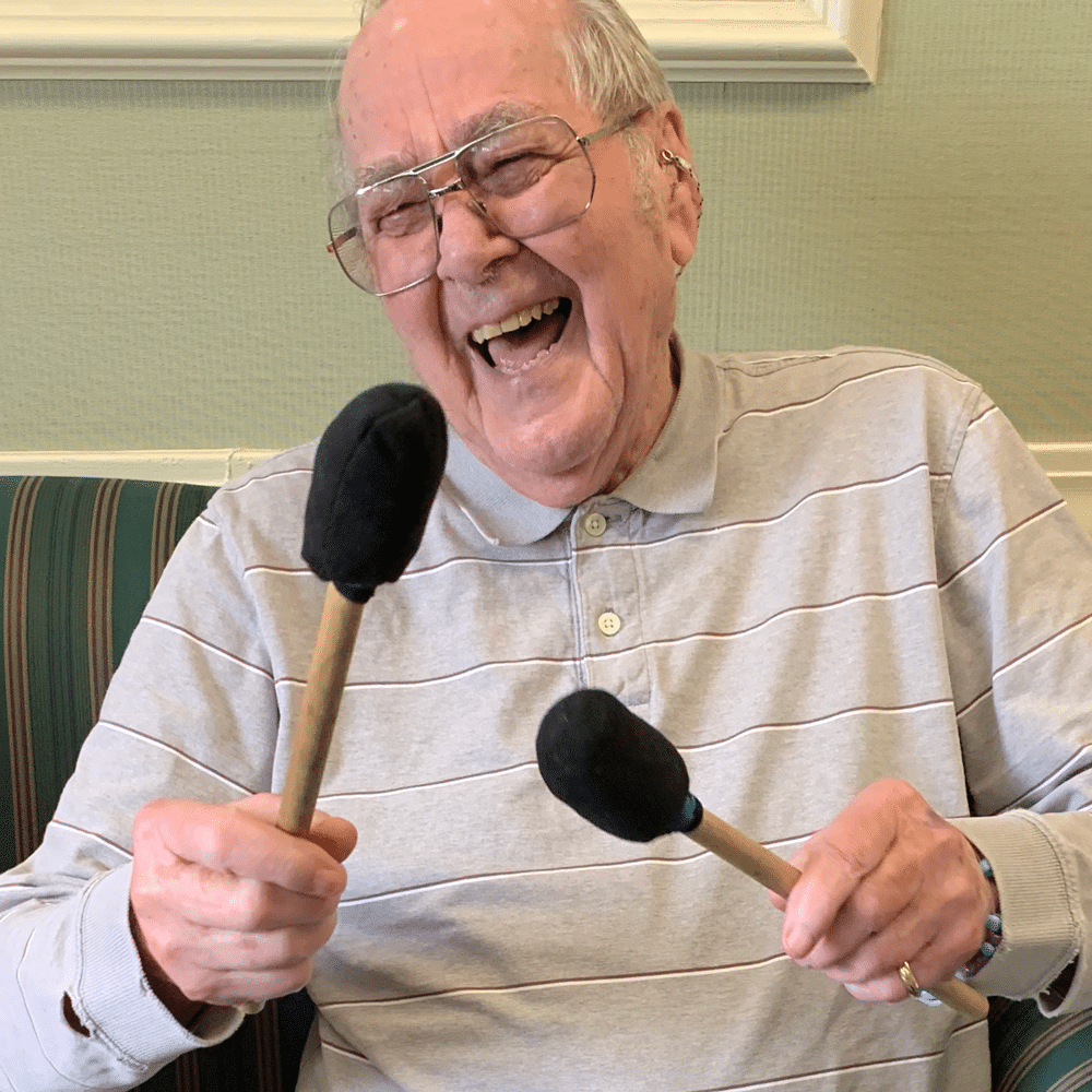 An elder man smiles joyfully while holding two mallets and playing a drum