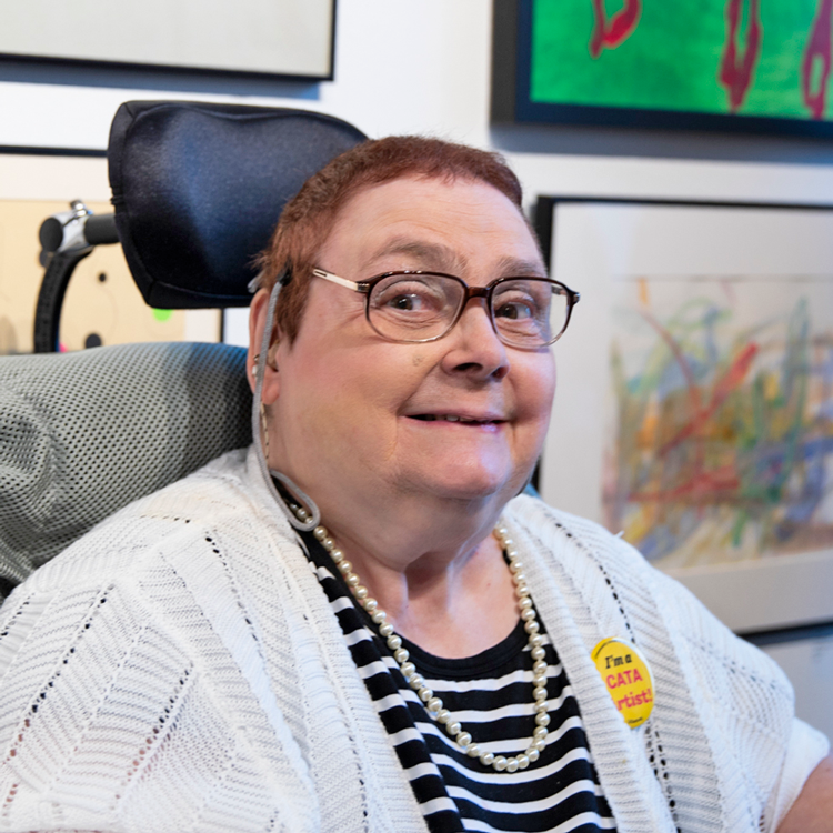 White woman in a wheelchair wearing a black and white shirt, white sweater, pearls, glasses, and smiling.