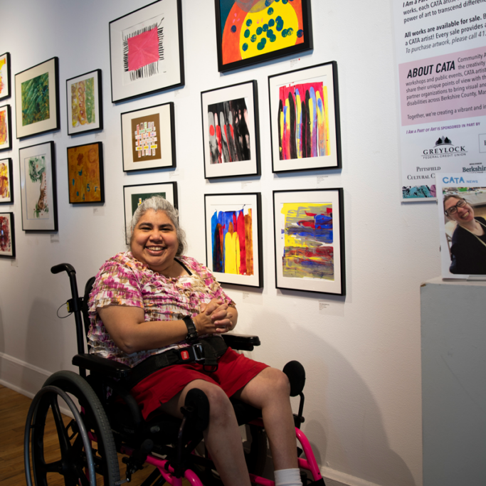 Connie in a wheelchair wearing a pink and white blouse, in front of a wall of paintings.