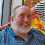 A white man with grey hair and a beard, smiling, wearing a blue and white striped shirt.