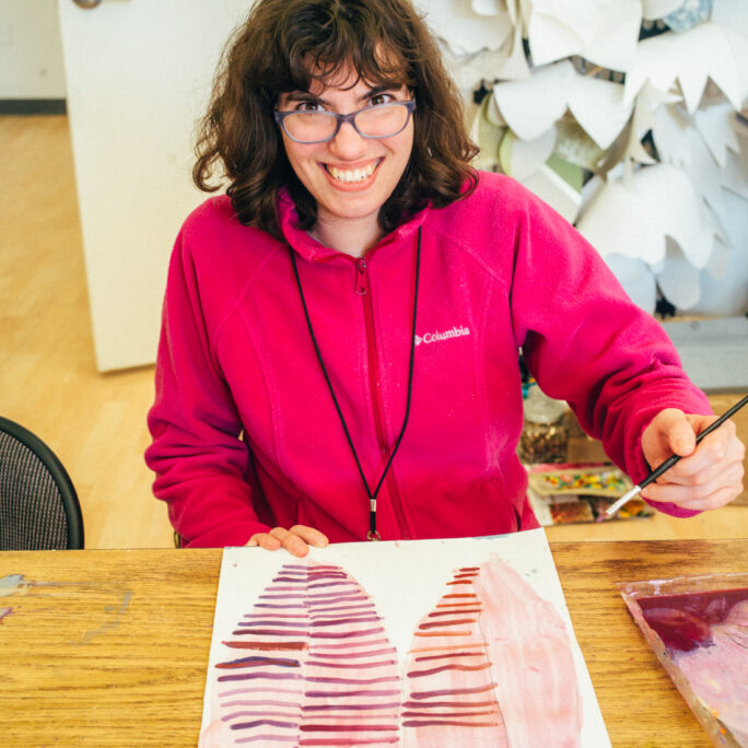 A woman in a pink jacket holds a paintbrush and smiles