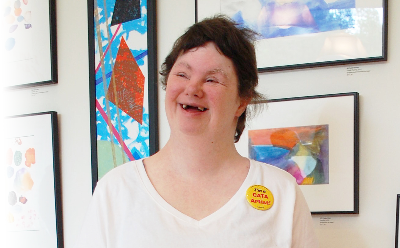 A woman with Down syndrome smiles in front of a wall of paintings.
