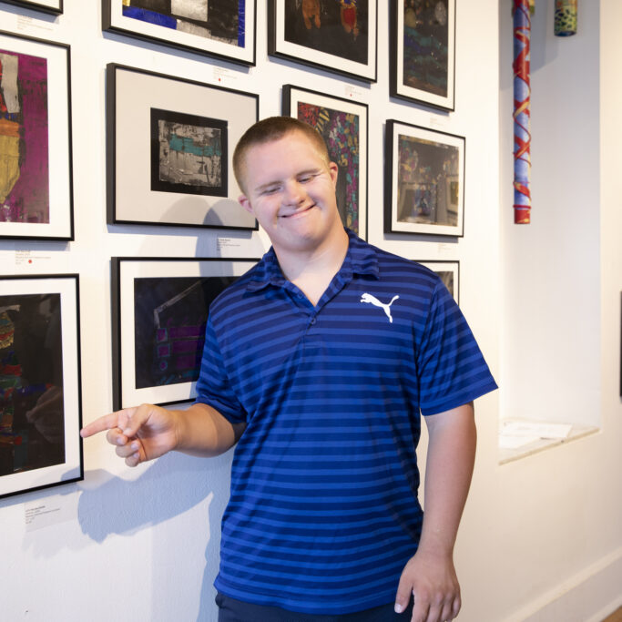 Image description: CATA artist Hayden Robb stands in an art gallery, smiling and pointing to his painting.