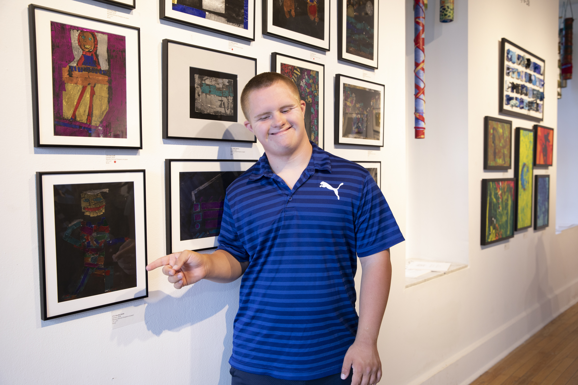 Image description: CATA artist Hayden Robb stands in an art gallery, smiling and pointing to his painting.