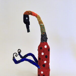 Mixed media bottle sculpture reminiscent of a bird. Creature has red painted fabric around the body with navy and white polka-dots. Two navy painted legs stick out of the body with curled bottoms created with pipe-cleaners. Creature has a long curved neck with a black beak-like head with a painted eye on the side. Creature's neck is covered with variously patterned fabrics.