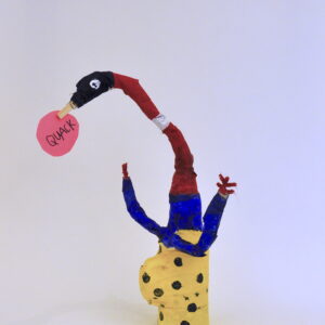 Mixed-media bottle sculpture reminiscent of a bird. Creature has a yellow body with navy polka-dots and a bulging stomach. Creature has two navy arms with red hands. Base of the creature's neck is navy blue, then becomes red and has a black head with a googly eye on the left side of its face. A clothespin beak holds a pink post-it sign that says "QUACK".