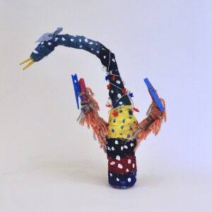Mixed-media bottle sculpture resembling a bird with a painted navy body and neck covered in white polk-dots. Middle of stomach has a red paint stripe, and yellow paint with blue polka-dots. Creature has two arms wrapped in orange and brown yarn, and two blue clothespins as the talons. Head has a drawn, black eye, a clothespin as a beak, and a blue-fabric headband. A wire with blue and red stars drapes around the creature's neck.
