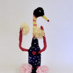 Mixed media bottle sculpture resembling a duck. Creature has navy painted fabric around the body with white polka dots and a heart in the center, two light-pink fabric roses are glued on as feet, two red arms with tufts of yellow yarn as arms. The creature's neck is painted with horizontal stripes of yellow and pink, it has a black head with a googly eye and a yellow painted beak.