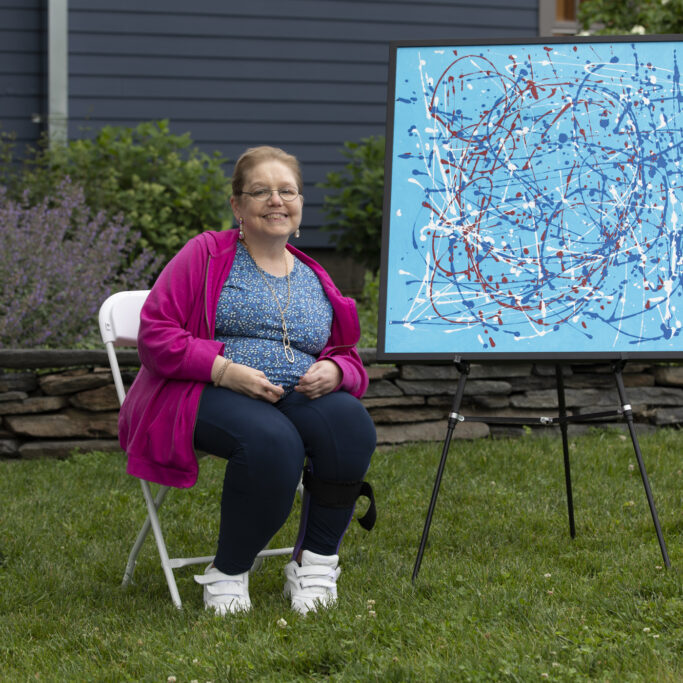 Betsy sits outside in a pink coat smiling next to her large framed painting on an easel