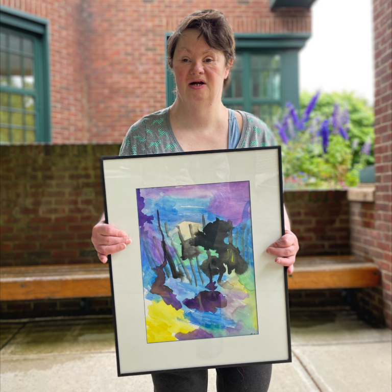 Image description: Cathy stands outside and holds her framed painting
