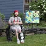 Chris sits in a wheelchair next to his painting on an easel