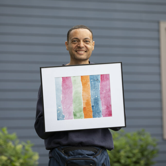 Franck stands outside smiling and holding his framed painting