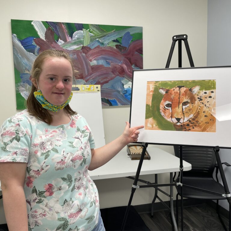 A young woman smiles and points to a framed painting of a cheetah