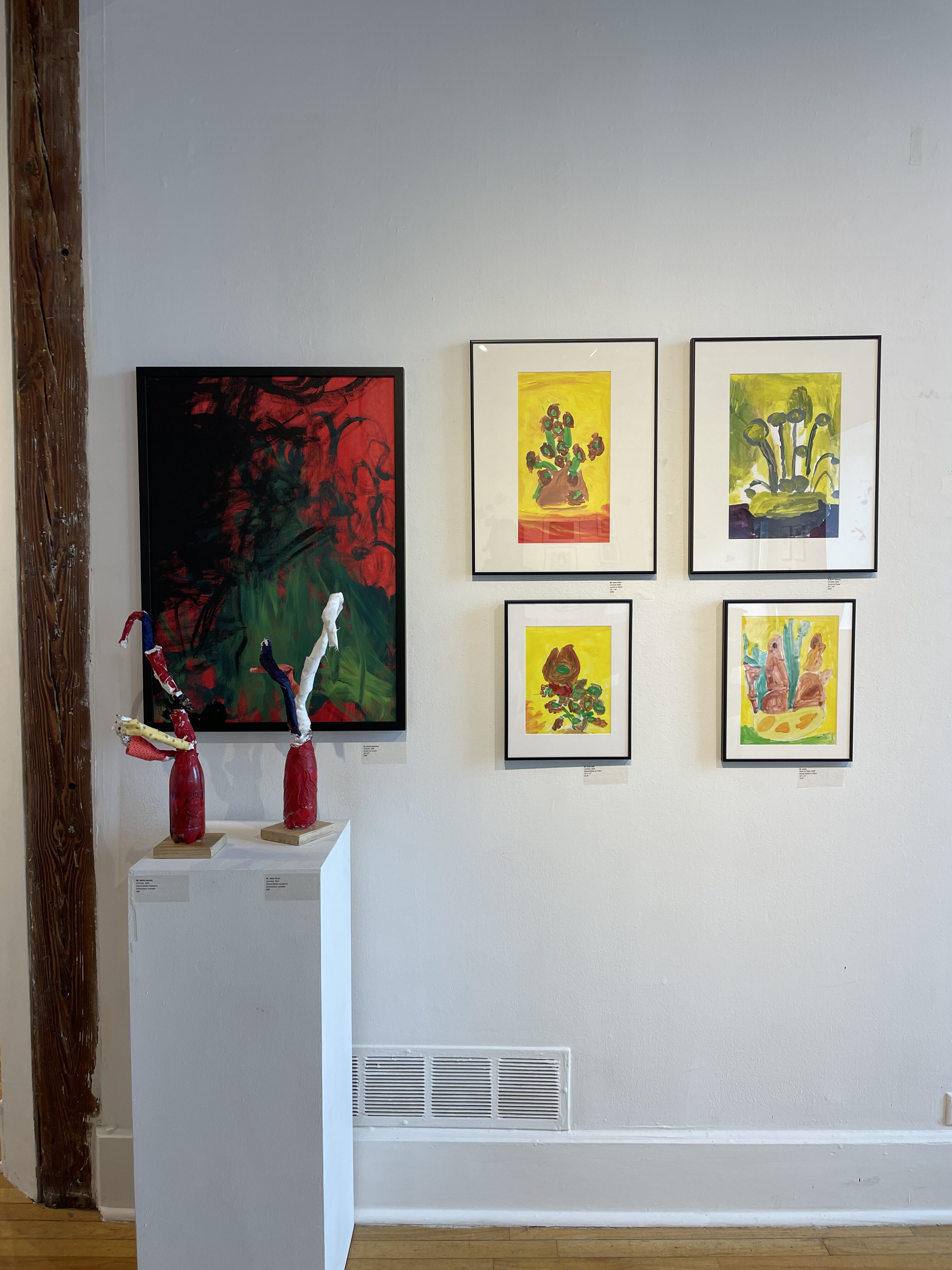 Gallery wall with a large red and green abstract painting alongside four small yellow and green paintings. Two sculptures sit on a white stand.