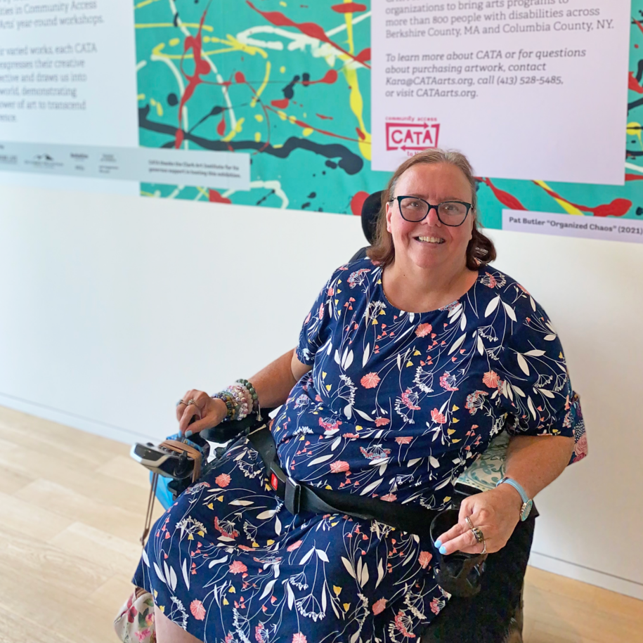 Image description: Pat in a wheelchair smiling in front of a large banner with her painting at the Clark
