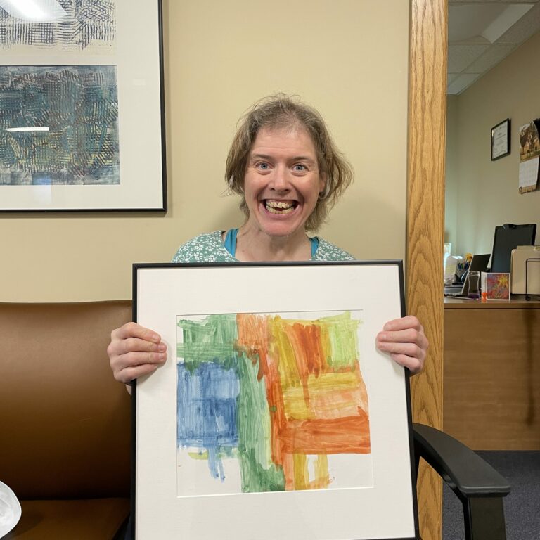 mage description: Joanne sits and smiles while holding her framed painting