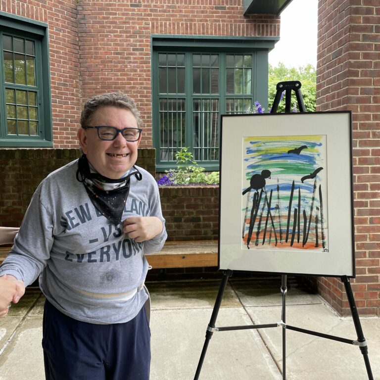 Image description: Joe stands outside smiling next to his framed painting on an easel