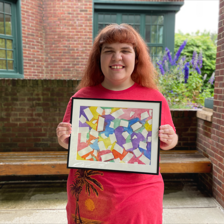 Image description: Karlie stands outside smiling and holding her framed painting