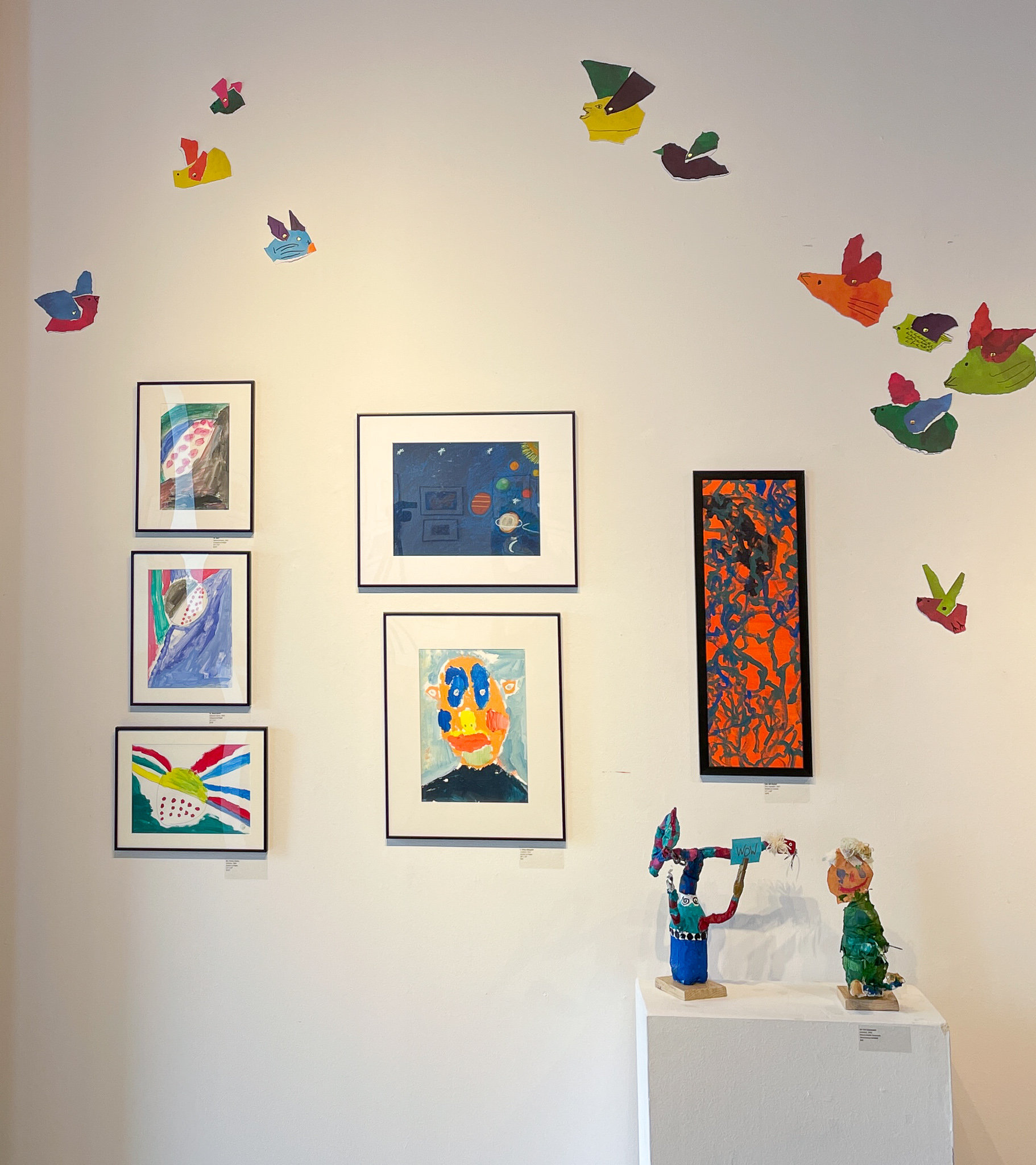 A gallery wall with six paintings, two sculptures, and an array of paper birds