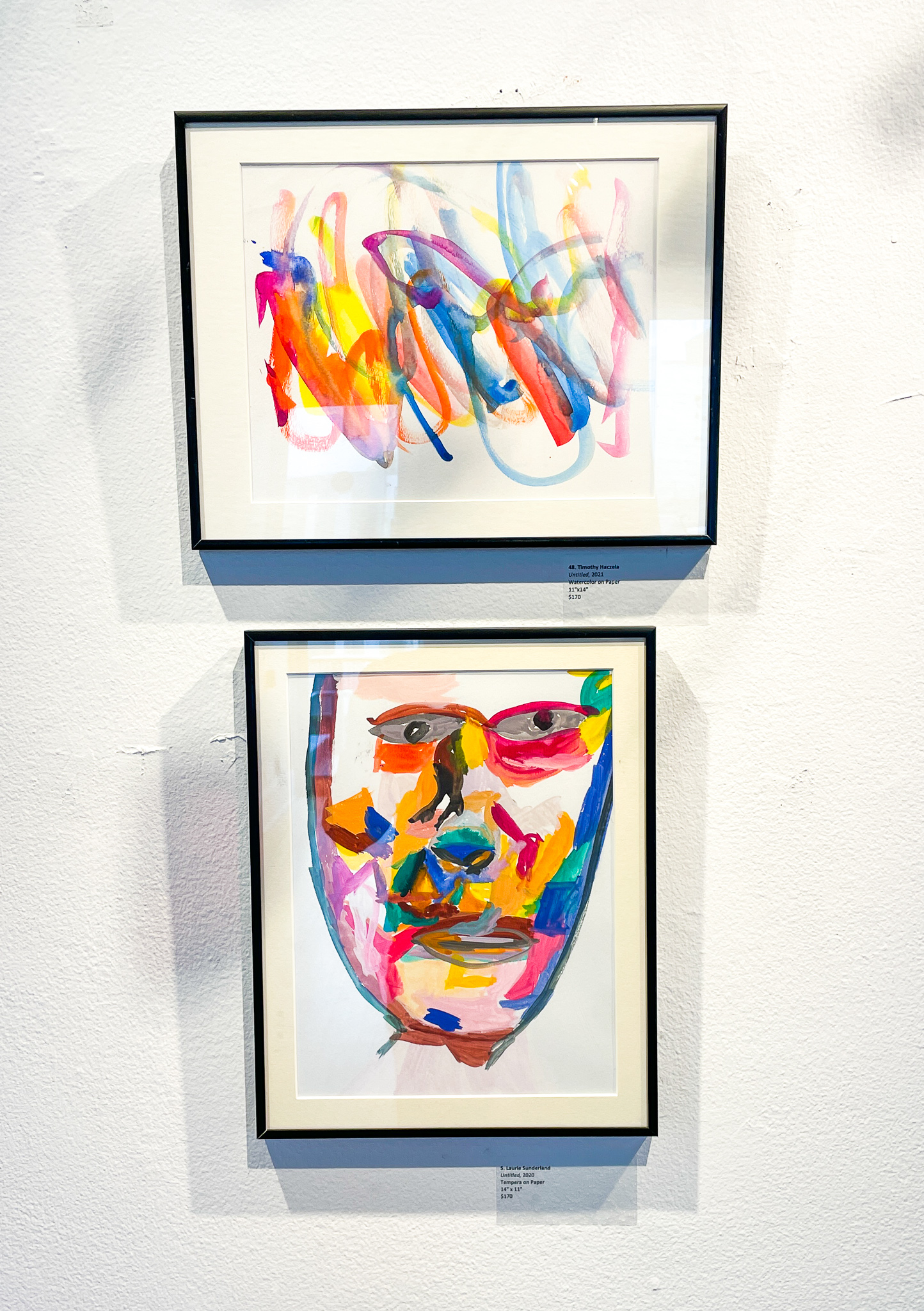 A gallery wall with two paintings: an abstract painting with multicolored brushstrokes on top and a multicolored portrait of a face on bottom