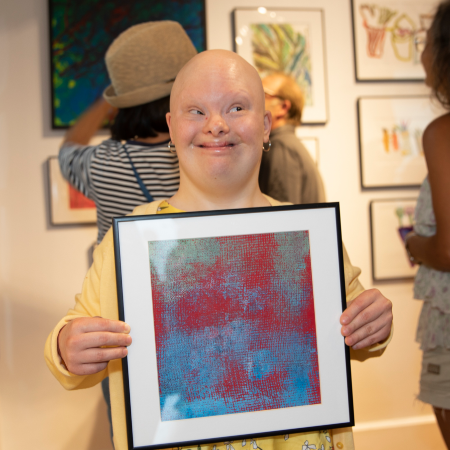 Louisa stands smiling and holding a framed painting in an art gallery during CATA's 2019 Annual Art Show