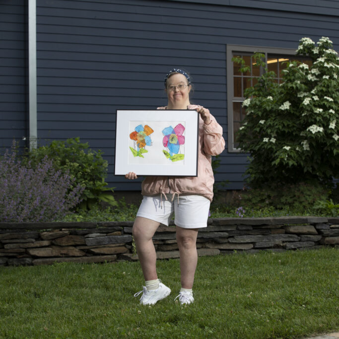 Image description: Margaret stands outside smiling and holding her framed painting