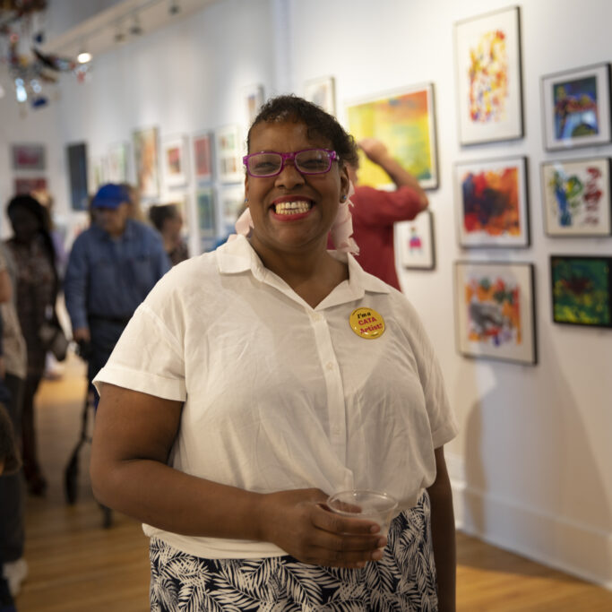 Image description: Natasha smiles and stands in an art gallery during CATA's 2019 Annual Art Show
