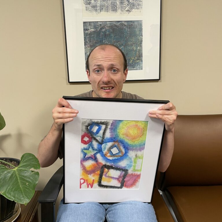 Image description: Peter sits smiling and holding his framed painting