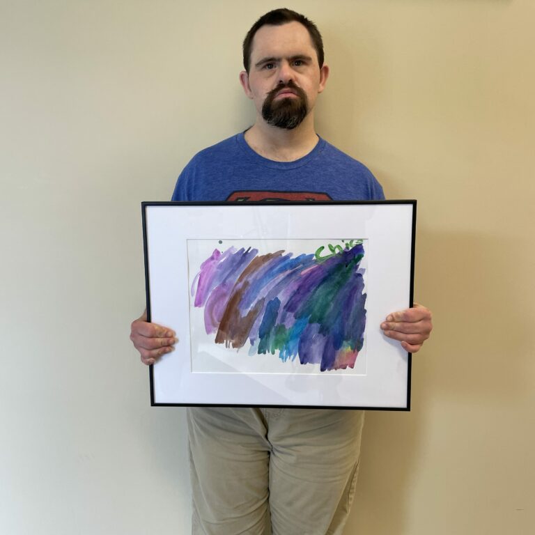 Image description: Ralph stands against a beige wall and holds his framed painting