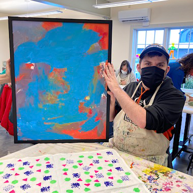 Image description: Artist TJ sits in an art studio holding his framed, orange and blue abstract painting