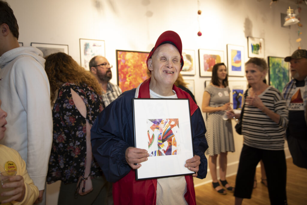 Image description: CATA artist smiling and holding a framed painting in a crowded art gallery