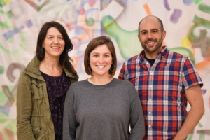 CATA co-Program Directors Kara Smith, Kelly Galvin, and Jeff Gagnon smile together in front of a colorful, graffiti-style backdrop.