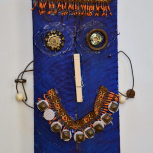 A face on a piece of blue painted wood. There is a beaded necklace as the hair and mouth. A clothespin for a nose. And pieces of metal make up the eyes, part of the hair, and the ears.