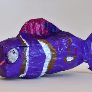 A purple long and thin paper Mache fish with a pink smile, big eyes, a pink fin, and black and white stripes.