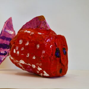 A paper mache fish painted red with white dots. The fish has purple eyes and a pink tail and fins. The tail has purple and white stripes.