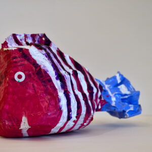 A paper mache fish with a pink head and white and purple stripes along its body. It has a light blue tail with a few white stripes.