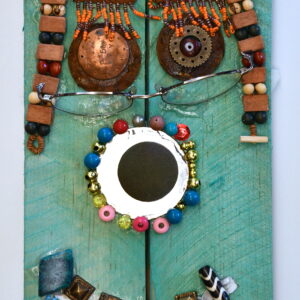 A face on a piece of Turquoise painted wood. There are metal pieces as the eyes eyes and pieces of beaded orange jewelry as the eyebrows. There is a beaded necklace as hair. A small circular mirror with beads around it as a nose. Blue beaded jewelry as a mouth with square pieces of metal, beads, and stones on top. The face is wearing glasses.
