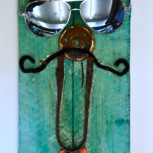 A piece of wood painted turquoise with objects glued on it that make a face. Circular metal pieces make eyes, nose, and mouth. Stones make the pupils. Beads are the eyebrows and teeth. There is a pipe cleaner as a mustache and the face is wearing sun glasses.