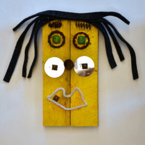 A face on a piece of yellow painted wood. There are metal pieces around green stones as the eyes and pieces of metal as the eyebrows. There are pieces of long rectangular fabric as hair. A small circular piece of metal is the nose and small circular shiny objects are cheeks. A white piece of pipe cleaner is made into a mouth shape at the bottom.