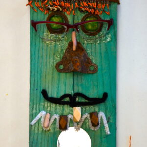 A portrait assemblage on wood that is painted turquoise. Orange beaded necklaces create hair and a beard. Spiky pieces of metal create some spiked hair. The face has glasses, green eyes made of stone surrounded by round pieces of metal. The nose is made of a big, rusted metal piece of hardware. Black pipe cleaner creates a mouth. And white pipe cleaner with stones and jewels create teeth. There is a small circular mirror beneath the mouth.