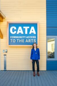Image description: Margaret Keller wears a blue shirt standing in front of a yellow wall and large blue and metal sign with CATA's name