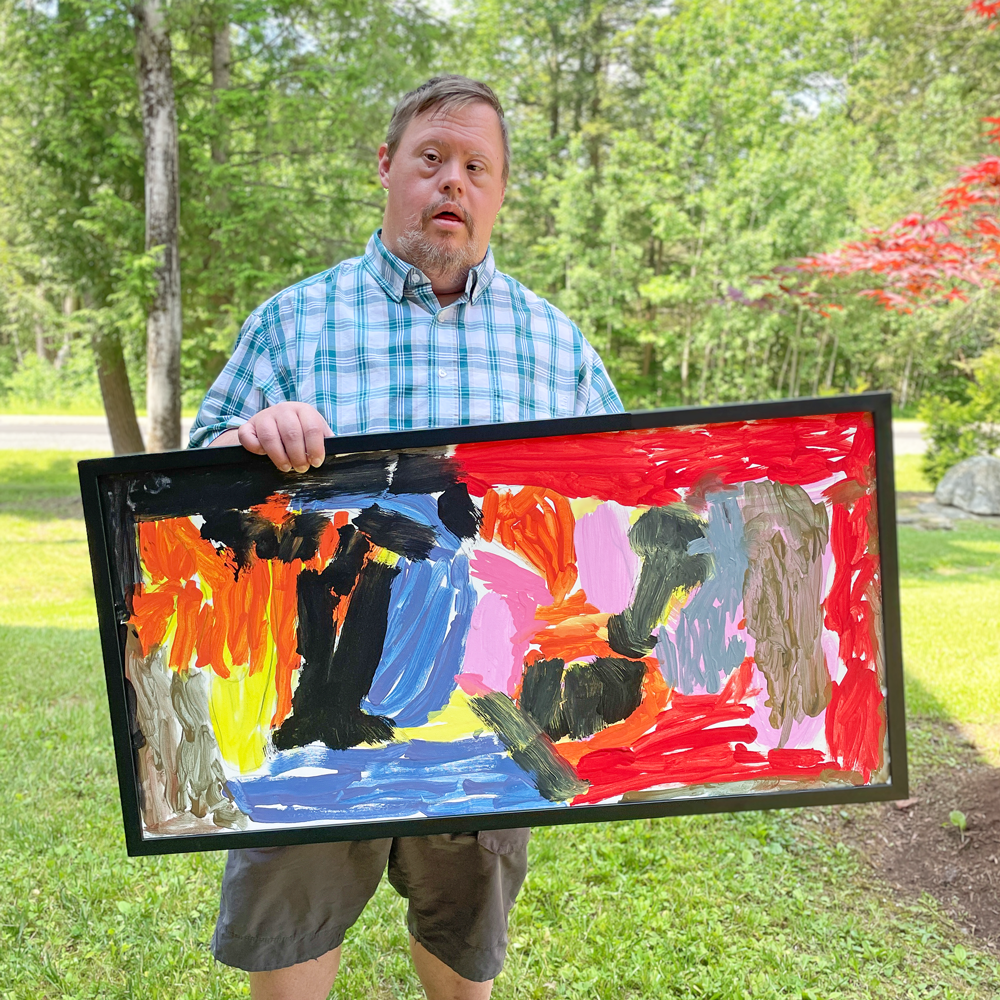 Image description: A man with Down Syndrome stands outside holding a large, framed abstract painting