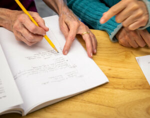 A Hand holding a pencil over a notepad while another hand points to the page