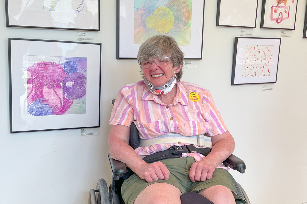 An artist in a wheelchair smiles in front of a wall of paintings