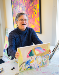 CATA artist Laurie Sunderland smiles and holds a finished painting of her family at the beach.