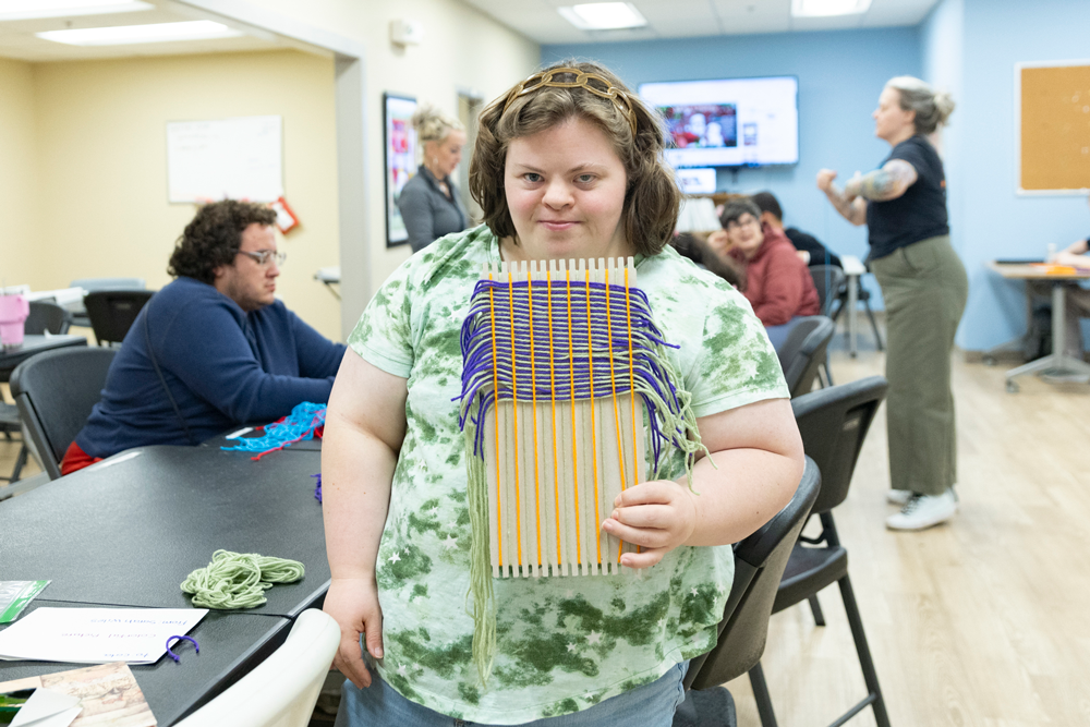 Sarah holds a purple and green weaving on a loom.