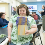 Sarah holds a purple and green weaving on a loom.