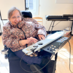 Terry smiles and plays a blue string instrument positioned on a stand in front of her.